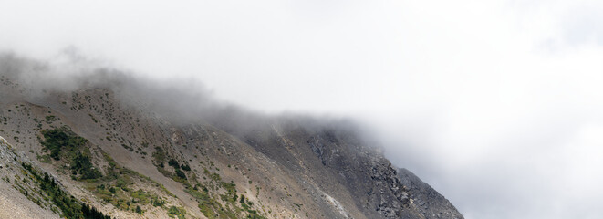 Panorama of mountain scene with scree and mist on top and partly transparent sky
