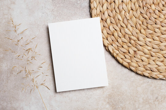 Blank invitation or greeting card mockup with botanical decor and natural straw placemat. Boho style flat lay with copy space for card decor