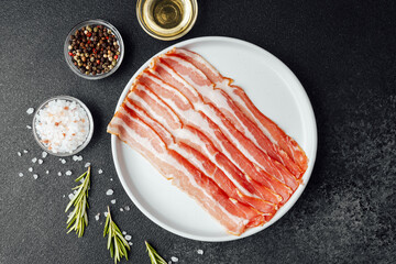 Raw bacon slices on white plate with pepper, salt and rosemary