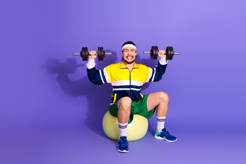 Active male athlete lifting dumbbells while seated on an exercise ball in retro sportswear against...