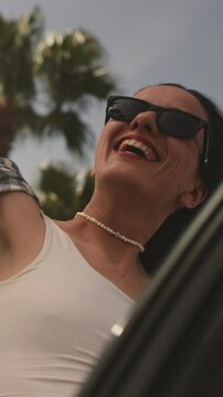 Vertical: Excited cheerful girl smiling. Female is enjoying a vacation in tropical city by sea or ocean. Woman in sunglasses, having fun on exotic island seaside road. Palm trees are reflected in car