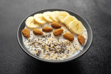 Oatmeal porridge with banana slices almonds and chia seeds in dark bowl on black background