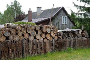 Pile of logs stacked behind wooden fence with  house in background in polish village, Poland