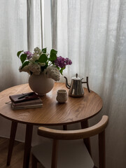 A bouquet of lilacs in a vase, a glass, a coffee pot, books on a round wooden table in the living room