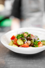 A vibrant and fresh Mediterranean salad in white bowl, featuring chopped cucumbers, tomatoes, red onions, olives, parsley, and crispy croutons. The blurred background enhances appetizing presentation