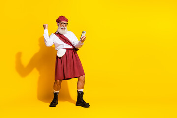 Cheerful senior man in Scottish attire holding phone celebrating on vibrant yellow background