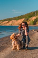 A curly-haired brown Labradoodle or Cavapoo dog with a young woman are best friends by the sea at sunset.