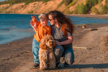 A curly-haired brown Labradoodle or Cavapoo dog with a little child girl and mom are best friends by the sea at sunset. Family trip with dog