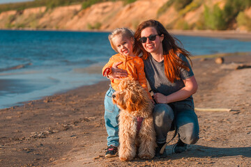 A curly-haired brown Labradoodle or Cavapoo dog with a little child girl and mom are best friends by the sea at sunset. Family trip with dog