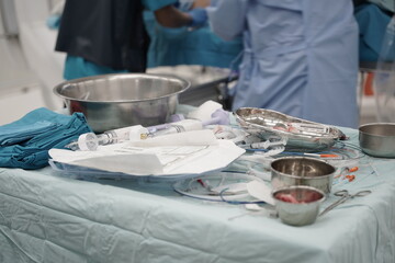 A surgical table holds syringes, metal bowls, and folded cloths, all neatly arranged for an operation. In the background, medical staff in blue gowns perform a procedure, focused and precise.