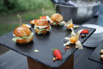 Elegant presentation of gourmet mini burgers on a slate tray, garnished with fresh strawberries and physalis. Ideal for upscale events, showcasing culinary artistry and premium ingredients.
