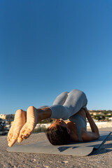 Fit woman practicing yoga shoulder stand outdoors on a mat near the ocean, under a clear blue sky, showcasing strength, balance, and mindfulness