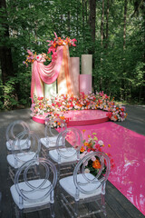 Outdoor wedding venue set up with a vibrant pink carpet leading to a stage adorned with colorful flowers. Transparent chairs line the aisle, creating a picturesque forest setting for a ceremony.
