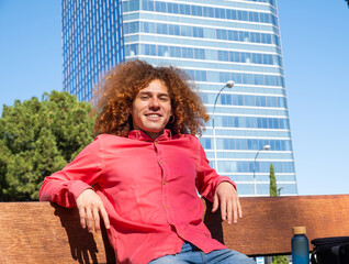Young man with curly hair relaxing on a bench in the city on a sunny day, wearing a red shirt, with modern building in background. Urban lifestyle concept