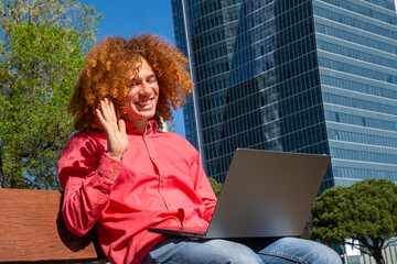 Side view oaf a young man with curly hair waving during a video call, working outdoors with laptop and reusable cup. Concept of remote work, green lifestyle and sustainability