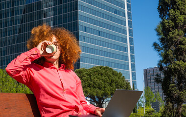 Young man with curly hair drinking from a reusable cup while working on a laptop outdoors. Concept of sustainable habits, remote work and eco-friendly urban lifestyle