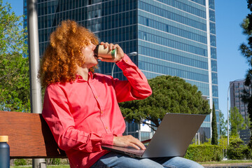 Young man with curly hair drinking from a reusable cup while typing on a laptop outdoors. Concept of sustainable remote work, eco habits and modern business lifestyle