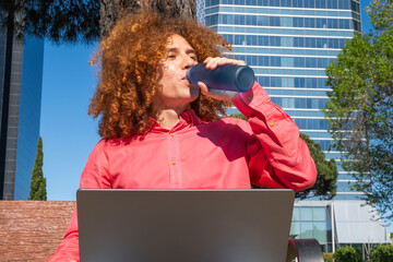 Front portrait of a Young man with curly red hair working on a laptop outdoors in the city, is drinking fresh water from a bottle near modern buildings. Remote work and digital nomad lifestyle concept