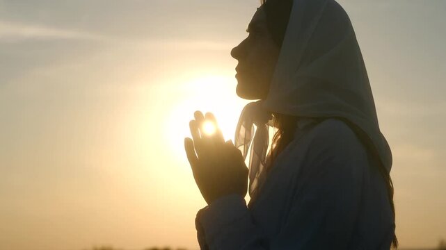 Golden hour sunlight illuminates woman in headscarf, her hands clasped in prayer, creating serene silhouette against colorful sky. Woman praying at sunset, concept of faith, hope. Young prayer woman.