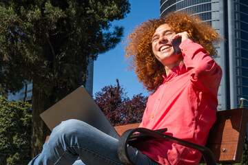 Cheerful young man with red curly hair having a phone call while using a laptop outdoors in a modern business district. Symbol of mobile work, eco-conscious living and digital connection