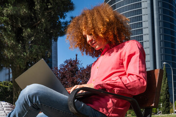 Confident young man with curly red hair sits on a bench working on his laptop in an urban setting. Modern office buildings behind him highlight the digital nomad and remote work lifestyle