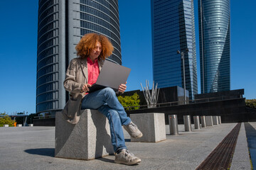 Young man with curly red hair working on a laptop in a modern financial district. He sits on a concrete bench with casual-smart outfit. Concept of remote work, focus and sustainable urban lifestyle