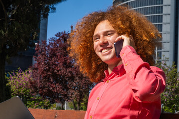 Close-up of a smiling young man with red curly hair talking on the phone, partially framed by a laptop lid. Modern remote work and sustainable lifestyle in an urban business setting