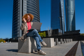 Young man with curly red hair waving during a video call, sitting on a concrete bench in a modern business district. Concept of remote communication, freelance work and sustainable lifestyle