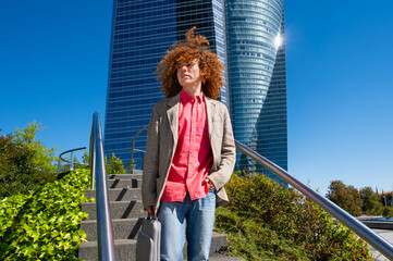 Young man with curly hair walking down stairs in a financial district, holding a laptop case. Modern office tower in background. Concept of green mobility, remote work and urban lifestyle