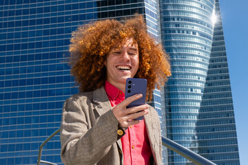Close-up of a young man with curly hair smiling at his phone in front of a modern office building. Concept of memes, social media, digital connection and contemporary urban lifestyle