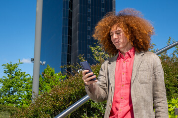 Young man with curly hair focused on his phone while sending a message in a financial district. He wears a blazer, with glass office buildings in the background. Urban freelance lifestyle