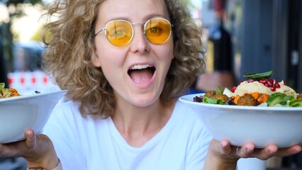 Excited curly blonde haired woman wearing yellow sunglasses showing two bowls of healthy vegan...