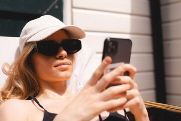 Calm cool blonde woman sitting and tanning on backdoor house, wear sunglasses, cap and swimsuit. Woman messaging, typing sms, read news, working online using mobile phone.