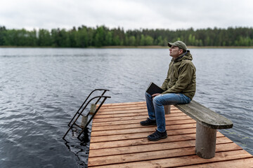 Man Sitting on Dock Holding Book, Looking Up Thoughtfully Over Calm Lake – Contemplation and...