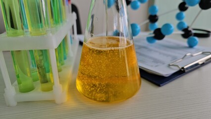 Glass flask contains a yellow liquid, bubbling slightly, alongside test tubes filled green liquid and a molecular model