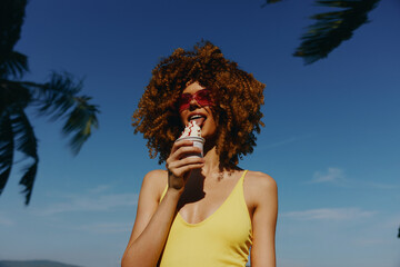 Young woman enjoying ice cream in the sun, wearing sunglasses and a yellow swimsuit The vibrant...