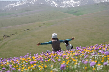 Happy woman with dog sitting on a field of flowers against the backdrop of snow capped mountains, rear view. Asy Turgen plateau in Almaty region of Kazakhstan in spring.