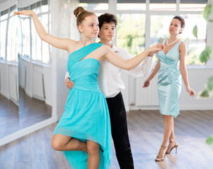 Motivated adolescent ballroom dancers, girl and boy in performance outfit practicing elegant dance moves in pair in bright studio, with female coach supervising in background