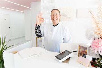 Smiling beautician making ok sign behind reception desk in beauty salon, showing satisfaction and excellent service. Looking at the camera
