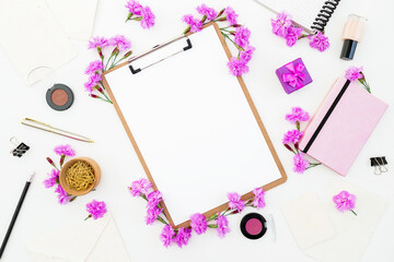 Workspace flat lay with clipboard, purple flowers, notebook, makeup and stationery on white background.