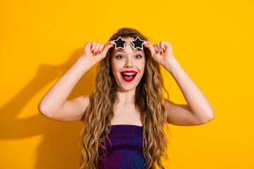 Playful young woman with curly hair poses with star-shaped glasses against a bright yellow background in a fun mood