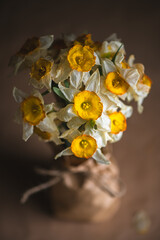 Bouquet of withering daffodils, their once vibrant yellow and white petals now fading and curling, sits in a rustic vase wrapped in burlap, evoking a sense of melancholy and the passage of time