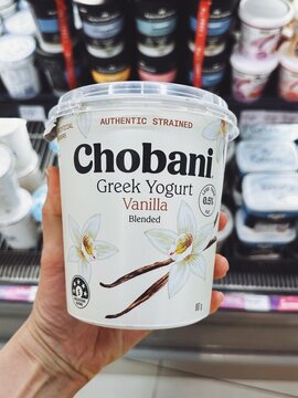 Large tub of Chobani Greek Yogurt in vanilla flavor, featuring clean white packaging with vanilla flower graphics. Held by hand in refrigerated section. Malaysia, Kuala Lumpur- May, 2025