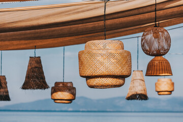 Wicker lamps of various shapes hanging from a wooden structure providing shade on a sunny day with...