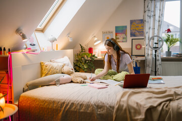 A young woman uses a laptop and writes in a notebook while sitting on her bed at home. Headphones on her head.