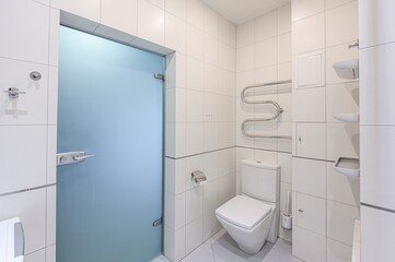 modern bathroom featuring a blue glass door, white tiles, toilet, and towel rack. Clean, minimalist aesthetic