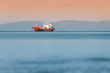 Cargo ship sailing on calm sea at sunset with mountains in background, maritime transport concept