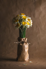 Wilting daffodils with yellow and white petals and green stems bundled in brown paper tied with twine, creating a melancholic still life against a brown backdrop