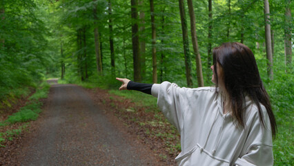 A young woman in a light beige hoodie in the forest pointing toward the road