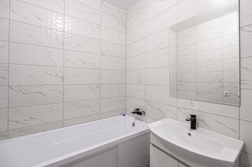 modern bathroom with white marble-like tiles, a white bathtub, a sleek sink with a black faucet, and a large mirror